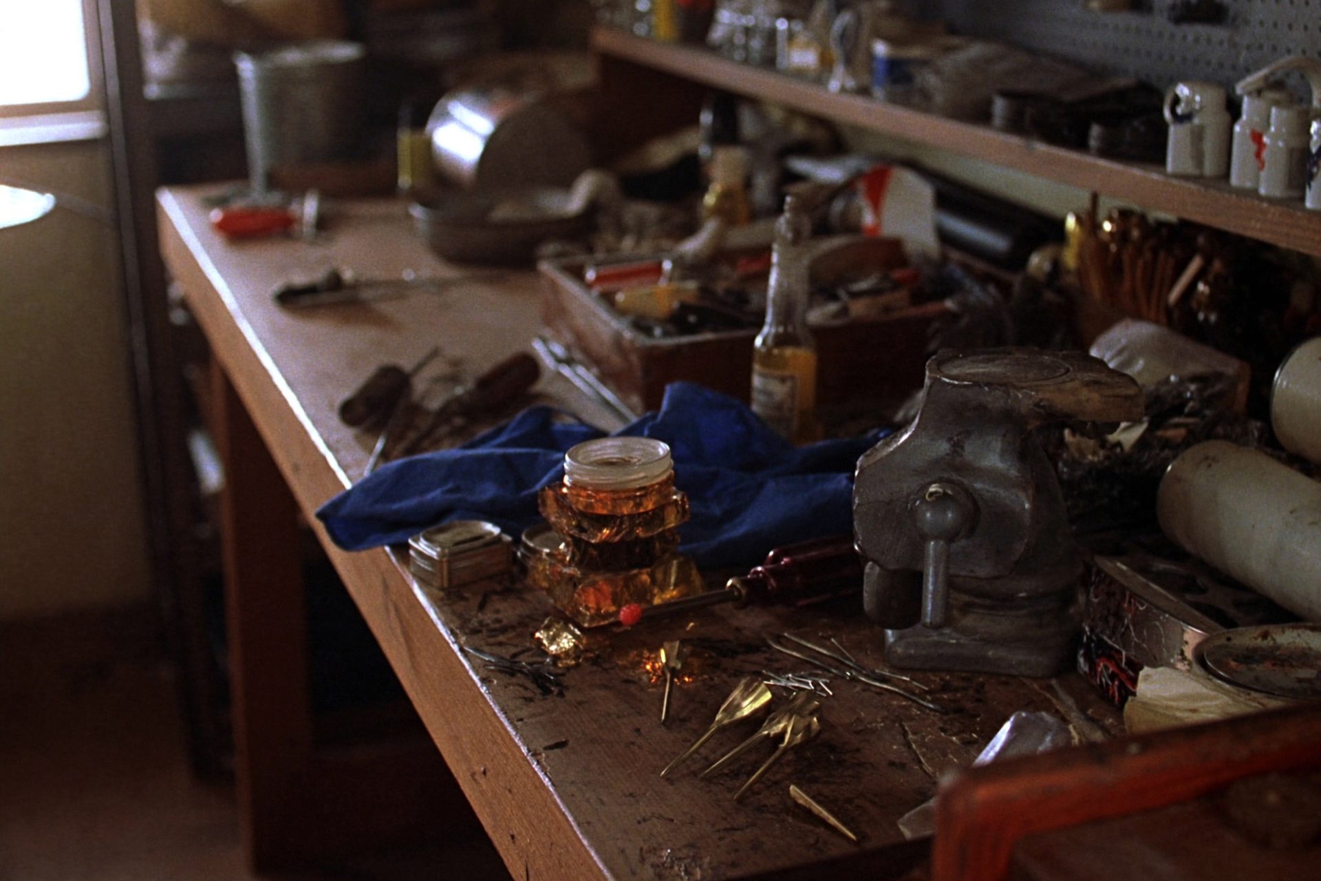 A gunsmith's workbench lit from a window on the left: a steel bench vise, brass punches, screwdriver bits laid out in a wooden tray, small parts in labelled tins and blue shop rags on a scuffed wooden benchtop.
