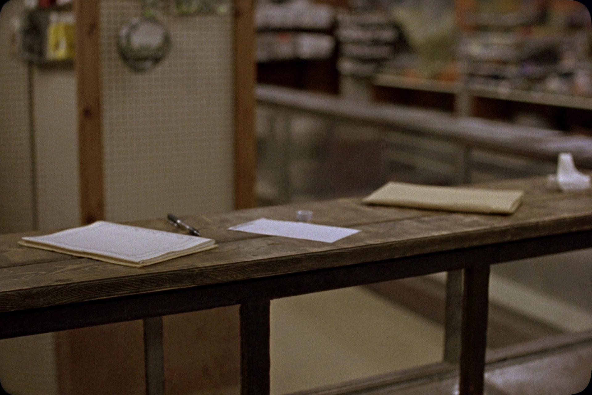 The wooden counter of a small rural Missouri shop: a glass display case with a worn top, an open ledger book and pen, a receipt spike and a cardboard shipping box set down beside them, a pegboard wall out of focus behind.